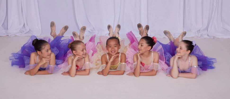 Five young JMDS ballet students in colourful tutus lying in a row, chins in hands