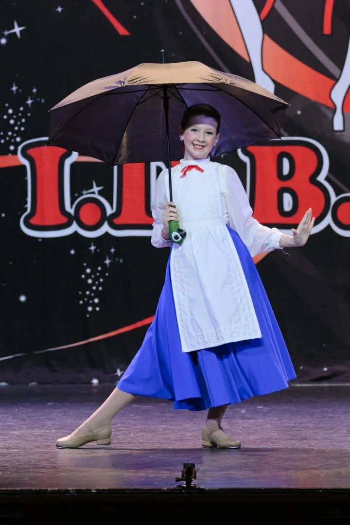 JMDS soloist on stage with an umbrella, in a blue skirt and white apron