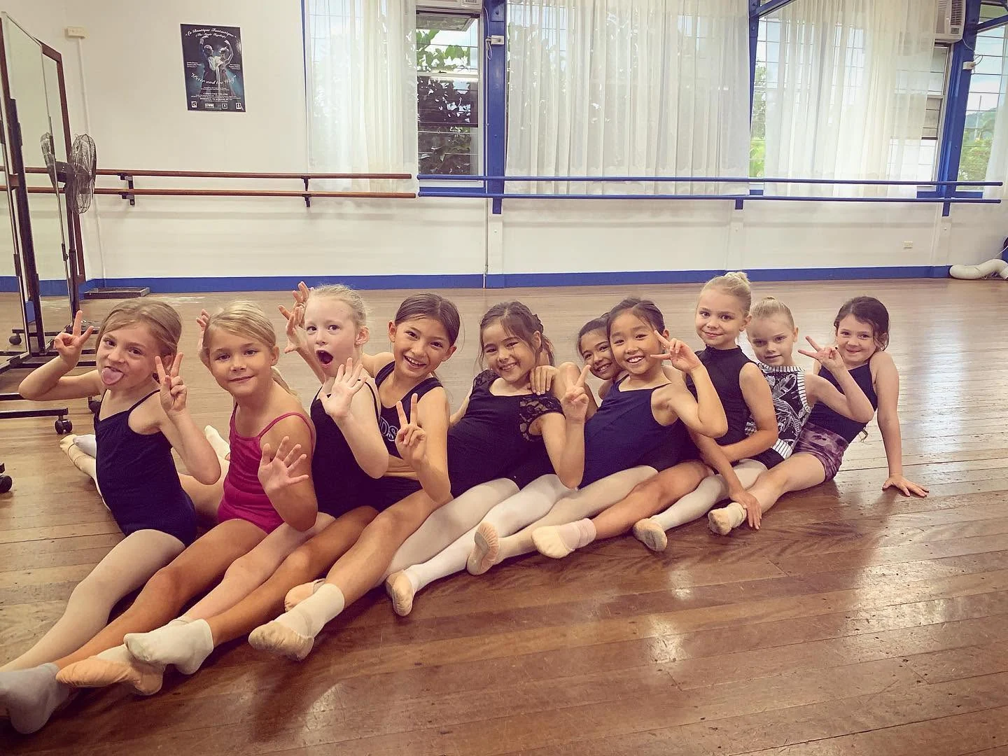 Young JMDS dancers sitting in a row on the studio's wooden floor, waving at the camera
