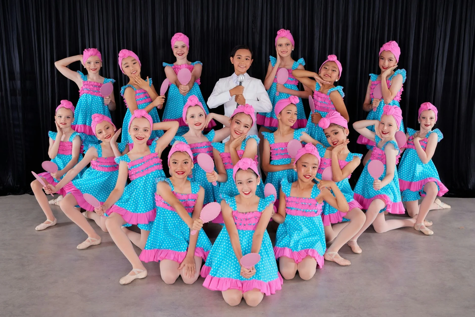 JMDS troupe in blue polka-dot dresses and pink turbans holding hand mirrors, around a dancer in a white suit